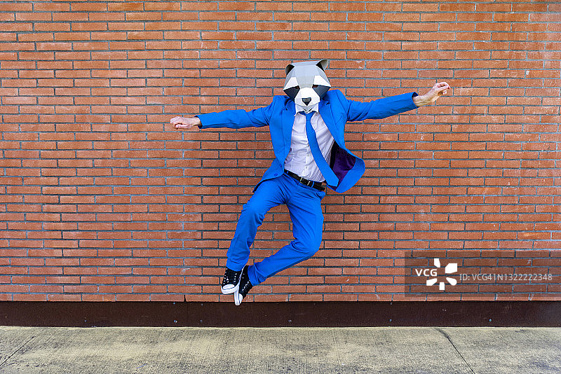 戴着蓝色西装和浣熊面具的男人跳跃在砖墙前图片素材