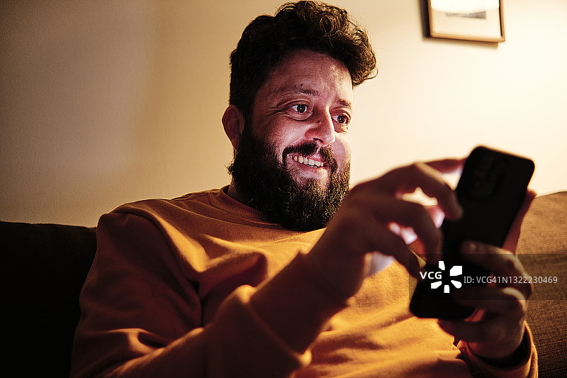 微笑的男人在家里的沙发上使用手机图片素材