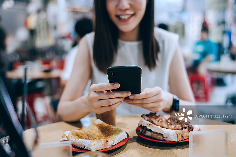 微笑的年轻亚裔女子在茶餐厅用餐时使用智能手机。她正在享用港式法式吐司和三明治，配上一杯传统奶茶。中华美食与饮食文化，生活方式与科技图片素材