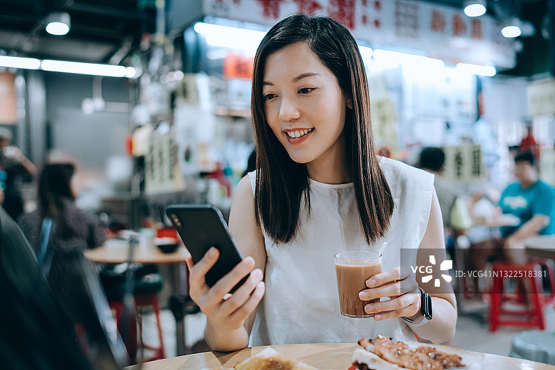 微笑的年轻亚裔女子在茶餐厅用餐时使用智能手机。她正在享用港式法式吐司和三明治，配上一杯传统奶茶。中华美食与饮食文化，生活方式与科技图片素材