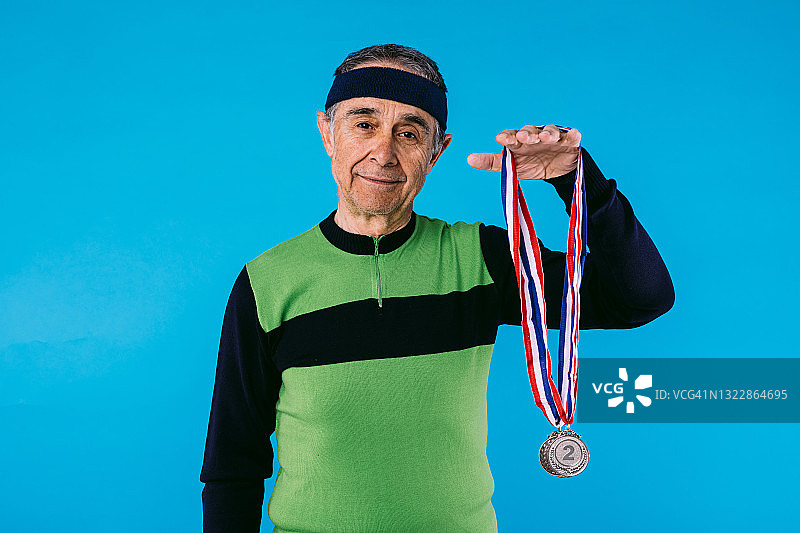 身穿绿黑色复古运动衫，手臂上挂着三枚奖牌的老年运动员，蓝色背景图片素材