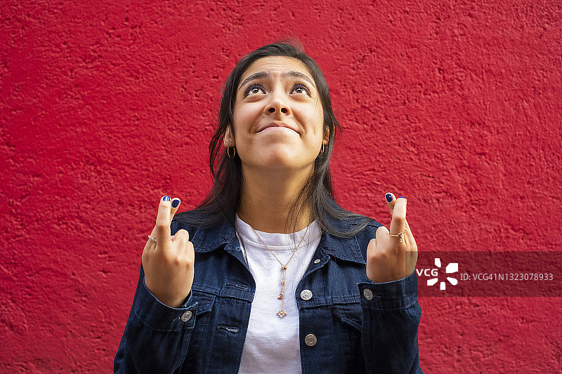 抬头，手指交叉，充满希望和祈祷的年轻拉丁裔女子，红色背景图片素材