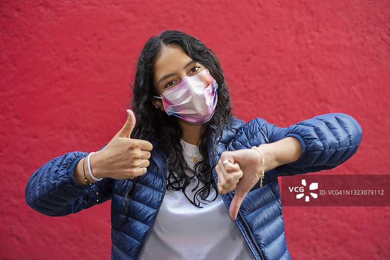 年轻拉丁裔女子戴着口罩，看着镜头，竖起大拇指，红色背景图片素材