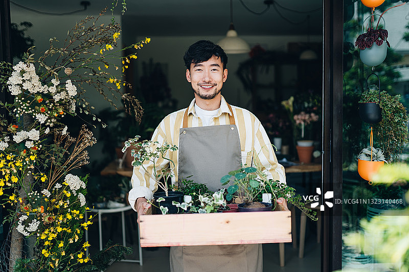 快乐的年轻亚洲男花店主，手捧鲜花木箱站在店外，面带微笑看向镜头，享受与花为伴的乐趣，小本生意概念图片素材
