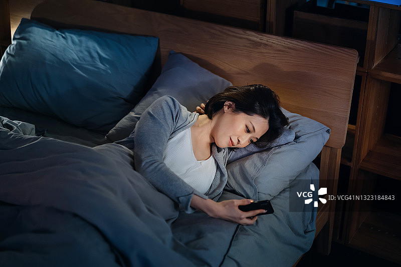 微笑的年轻亚洲女人在睡前躺在床上用智能手机查看社交媒体，在舒适的家中度过轻松的夜晚图片素材