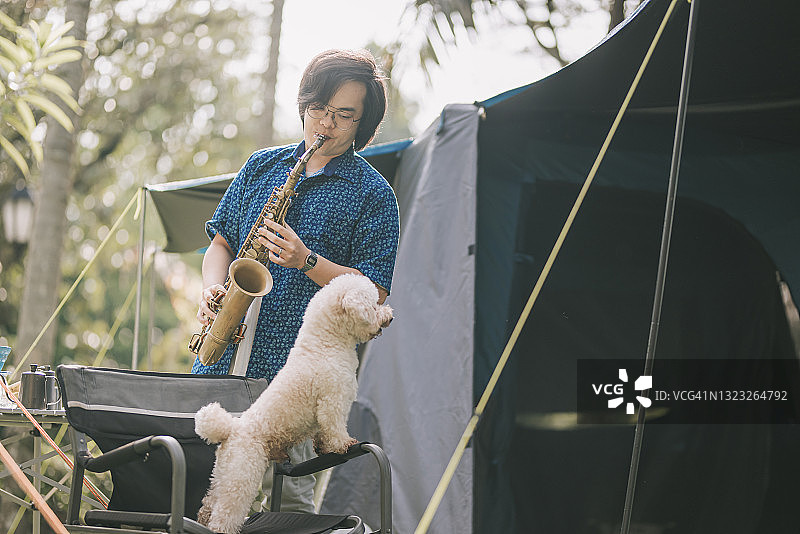 亚裔华裔萨克斯管演奏者清晨在露营帐篷前与宠物玩具贵宾犬一起演奏图片素材