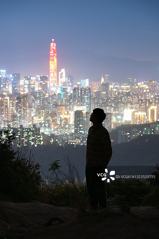 夜晚，在灯光璀璨、色彩缤纷的城市景观前，放松并仰望天空的男子轮廓图片素材