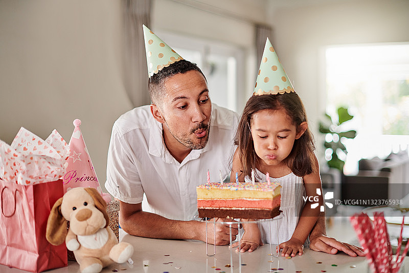 小女孩和父亲戴着派对帽吹灭生日蛋糕上的蜡烛图片素材