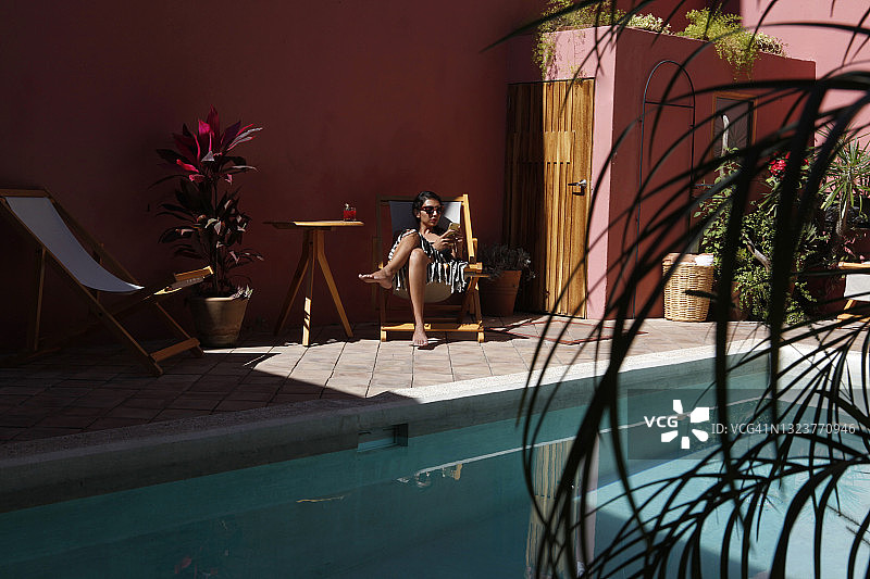 坐在游泳池边的椅子上，看手机的拉丁裔女人图片素材