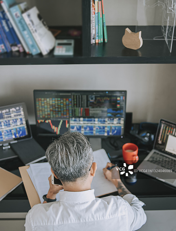 中国年长男性在家工作，通过多台电脑屏幕监控股市趋势（俯视图）图片素材