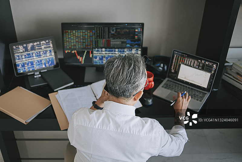 中国年长男性在家工作，通过多台电脑屏幕监控股市趋势（俯视图）图片素材