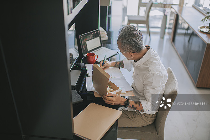 年长的亚洲男人在家工作，吃外卖，同时用多台电脑屏幕监控股市走势图片素材