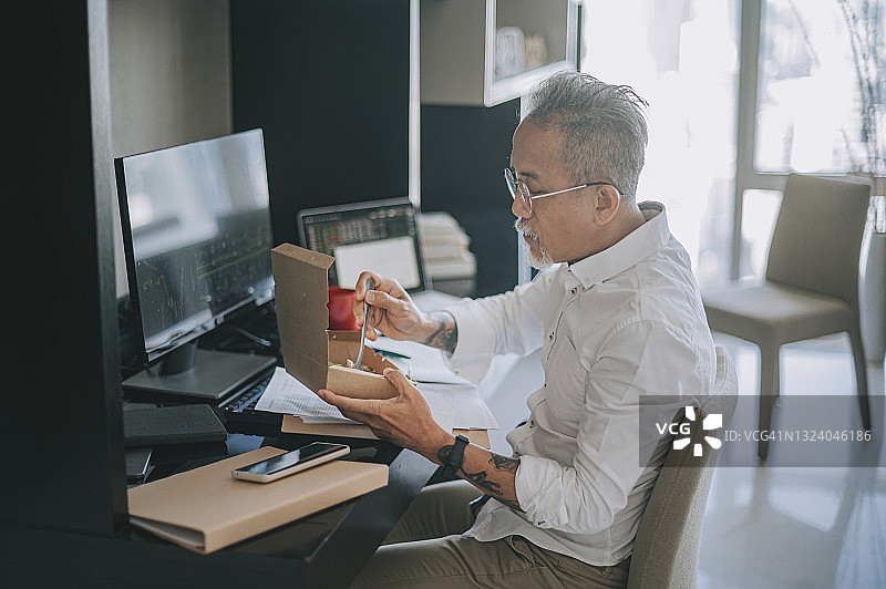 忙碌的亚洲老年 мужчина吃外卖，在家中通过多台电脑显示器监控股市走势图片素材