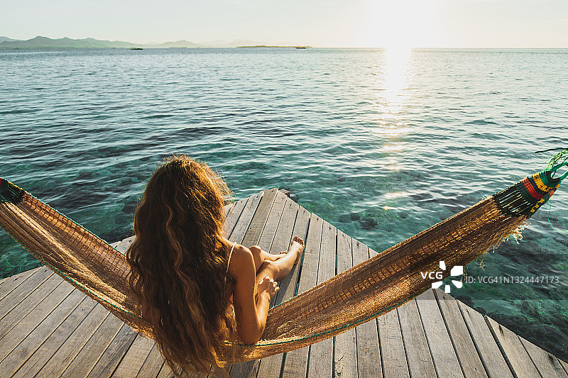 从后方看，女子在木制酒店露台上醒来，可欣赏海景，配有吊床和美丽的晨光图片素材