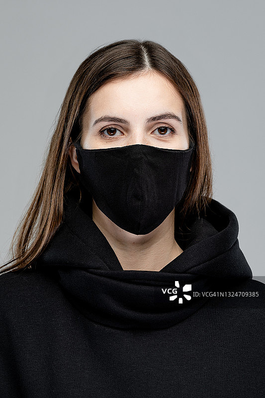 戴着医用口罩的现代女性在灰色背景的工作室里。高加索白人女士穿着黑色连帽衫图片素材