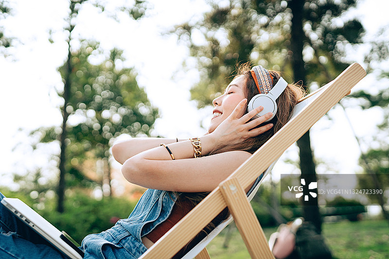 年轻漂亮的亚洲女性戴着耳机在公园躺椅上放松，享受夏日户外时光。音乐、青少年生活方式和科技图片素材