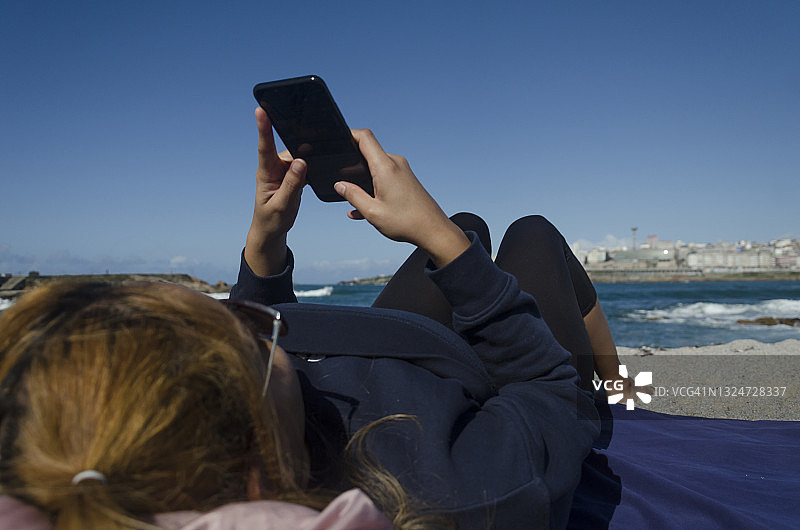 年轻女子在沙滩上看手机屏幕，西班牙科鲁尼亚城市背景图片素材