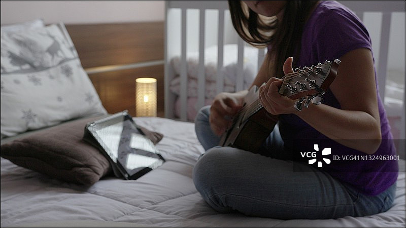 在卧室弹吉他，背景有婴儿床。新冠疫情期间在家用数位板进行吉他练习图片素材