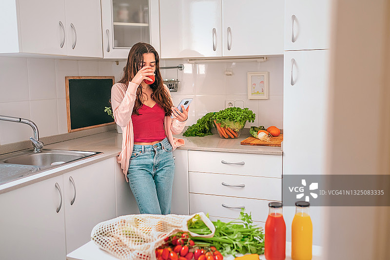 在厨房喝番茄汁和使用手机的健康年轻女人图片素材