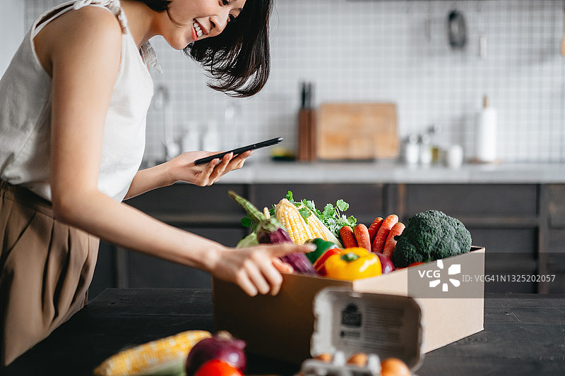 美丽的亚洲年轻微笑女性在家门口检查刚送达的装满从网上订购的色彩鲜艳的新鲜有机食品杂货的盒子，手里拿着智能手机图片素材
