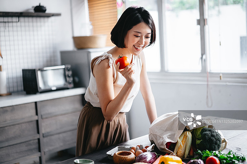 刚从超市购物回家的年轻亚洲女性，从可重复使用的购物袋中取出新鲜水果和蔬菜图片素材