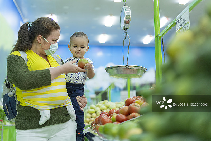 年轻的母亲有两个孩子，一个带着围巾的婴儿和另一个戴着口罩陪她去食品店买菜和蔬菜的婴儿图片素材