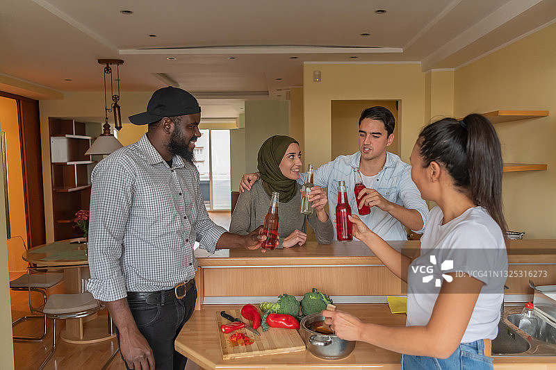 不同种族的快乐朋友在家里准备新鲜蔬菜晚餐，畅饮瓶装酒精饮料图片素材