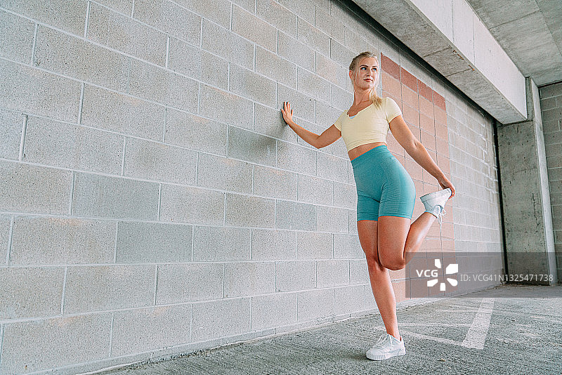 穿着运动服靠在砖墙上伸展四头肌的白人金发女人图片素材