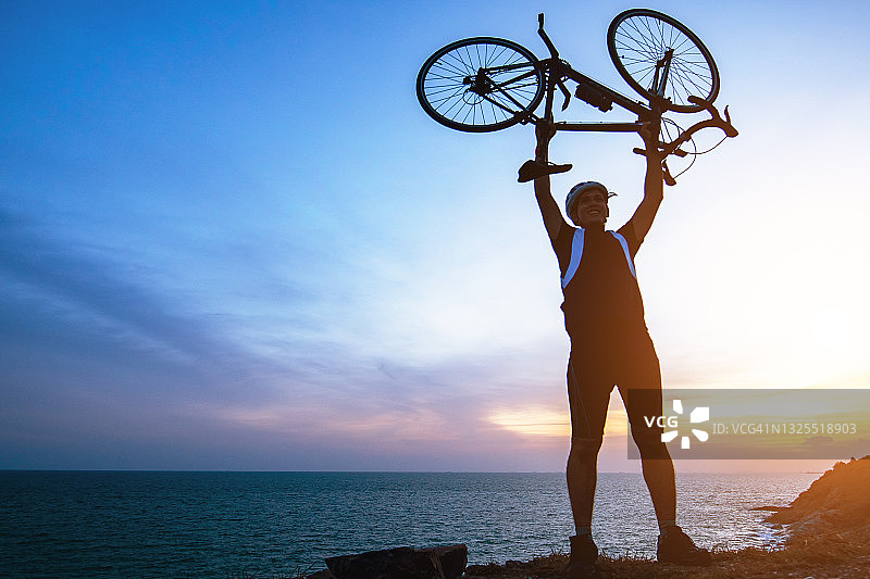骑自行车男子公路骑行，象征着成功、成就和胜利。快乐的男运动员在日落时分举起手臂，在海边举起自行车，欢呼并在山顶庆祝图片素材