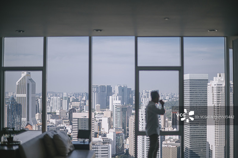 背影：中国籍亚洲退休老人清晨在客厅眺望吉隆坡首都城市，双手插在口袋里图片素材