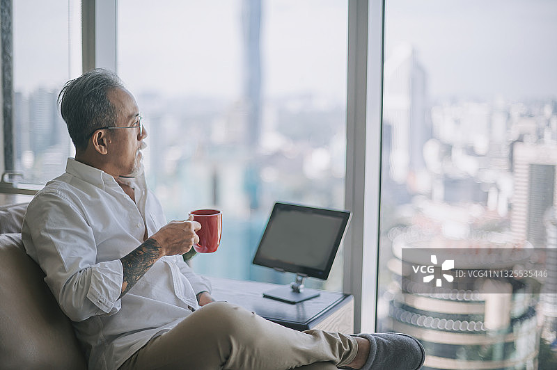 在客厅喝咖啡欣赏吉隆坡塔景色的华人老年男人图片素材