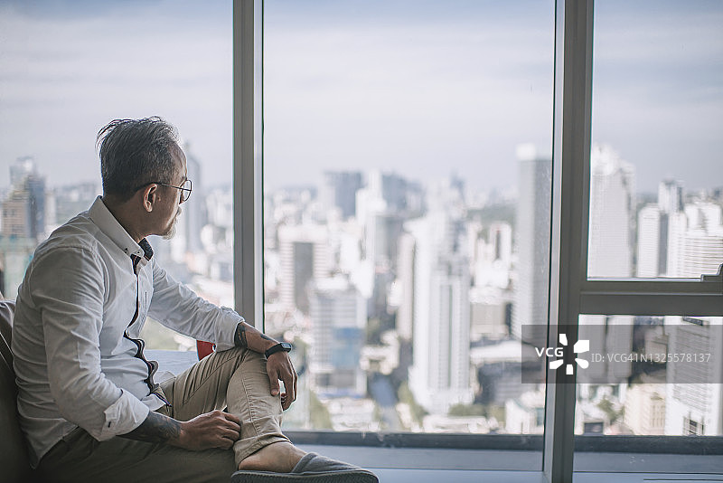 焦虑悲伤的亚裔中国老年男人白天坐在沙发上，透过窗户看着客厅图片素材