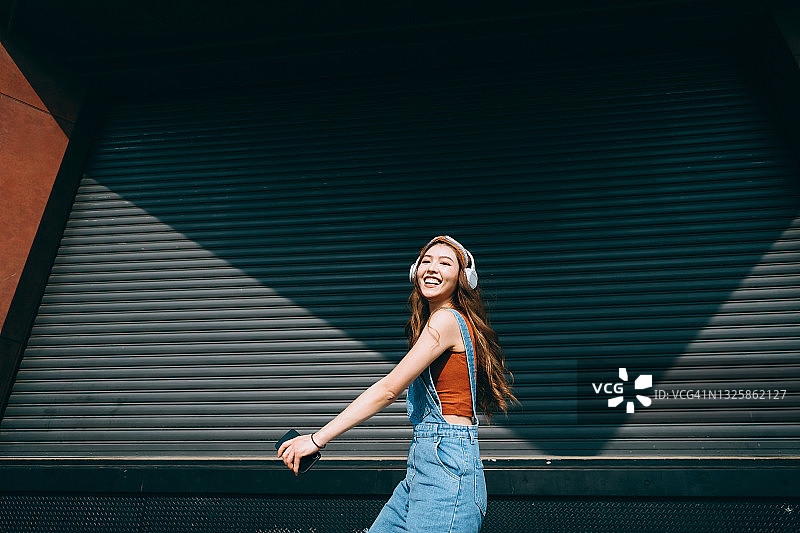 年轻亚洲女性户外彩色墙边戴耳机听音乐跳舞，手持智能手机图片素材
