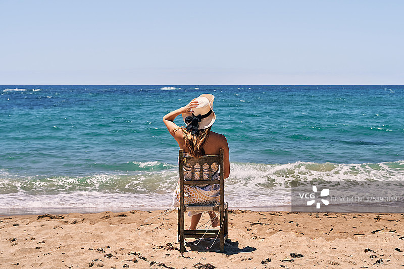 一位穿着泳衣和戴帽子的年轻女子坐在沙滩上的木椅上，手放在头上的后视图图片素材