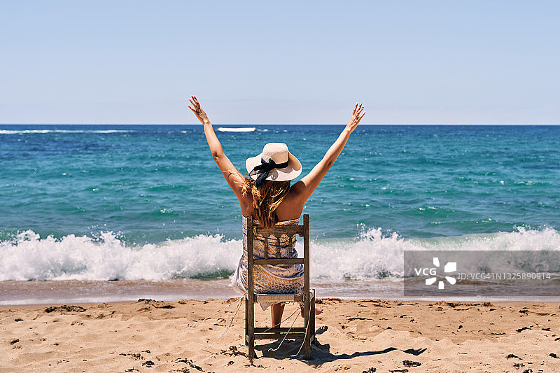 一位穿着泳衣和戴帽子的年轻女子坐在海滩木椅上，举起双臂的背影图片素材