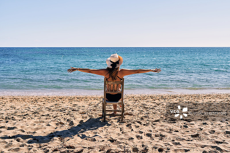 一位穿着泳衣和戴着帽子的年轻女子坐在沙滩上的木椅上，张开双臂的背面，无法辨认图片素材