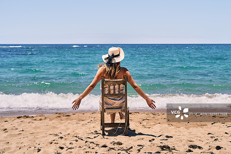 一位穿着泳衣和戴着帽子的年轻女子坐在沙滩上的木椅上，张开双臂的背面，无法辨认图片素材