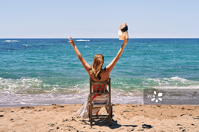 一位穿着泳衣和戴帽子的年轻女子坐在海滩木椅上，举起双臂的背影图片素材