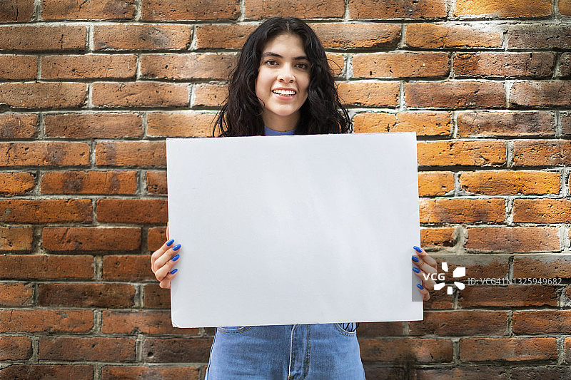 微笑并举着空白标牌的年轻拉丁裔女子，砖墙背景图片素材