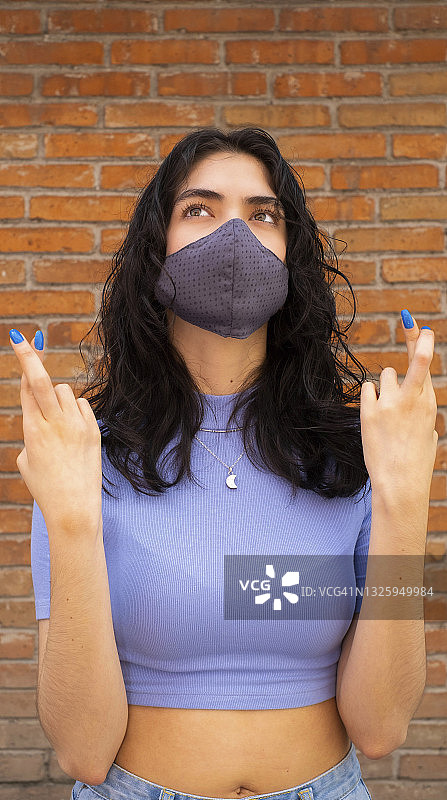 年轻的拉丁裔女人戴着口罩，抬头并交叉手指，充满希望地祈祷，砖墙背景图片素材