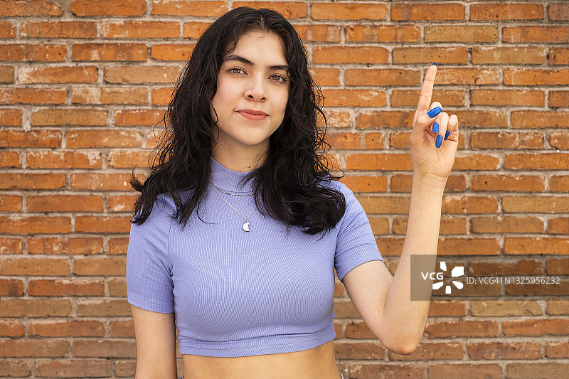 面带微笑，对着镜头做出第一名手势的年轻拉丁裔女性，砖墙背景图片素材
