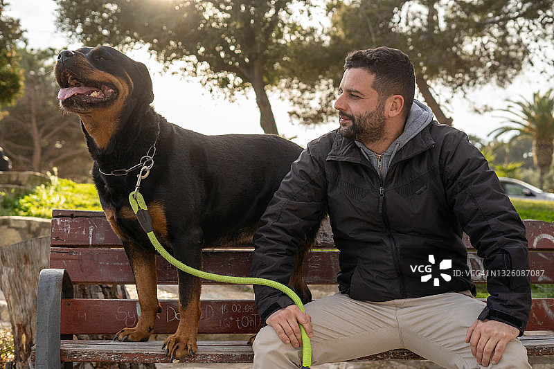 男人坐在公园长椅上，旁边是他的罗威纳犬图片素材
