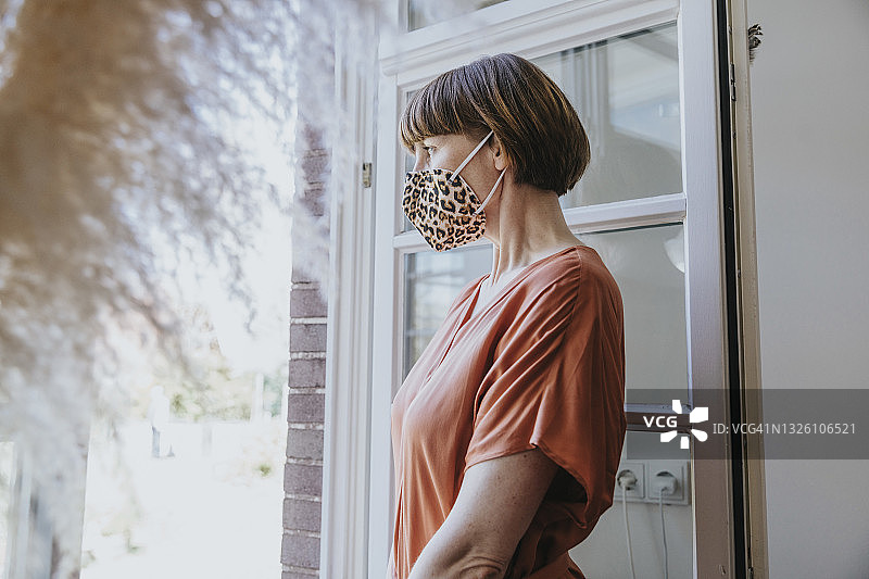 戴着动物纹口罩，在家中透过窗户向外看的女人图片素材