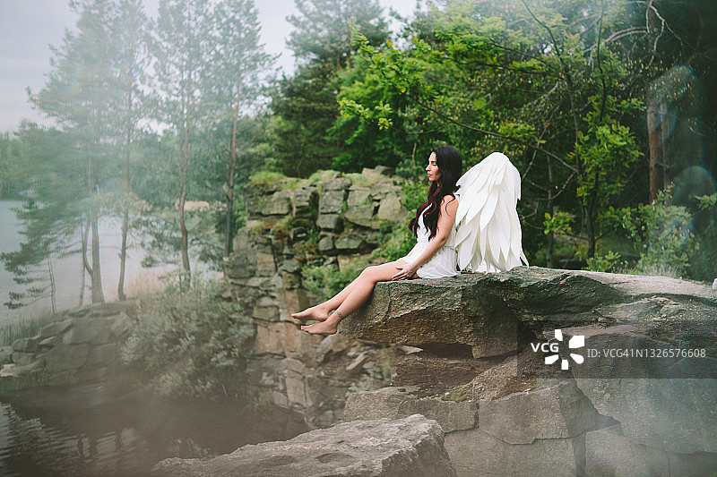 戴着天使翅膀、穿着白色连衣裙的深发模特坐在湖边采石场的悬崖上，背景是乌克兰的春天图片素材