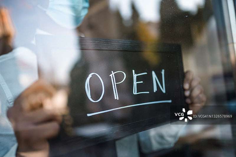 混血服务员在咖啡馆的窗户上拿着“营业中”的牌子，戴着口罩图片素材