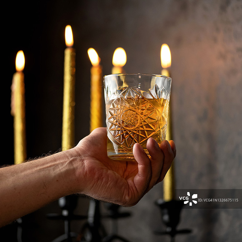 男性手持酒精饮品：男子在黑暗背景下，手持装有金色酒精饮品的水晶杯，点燃蜡烛，放松概念图片素材