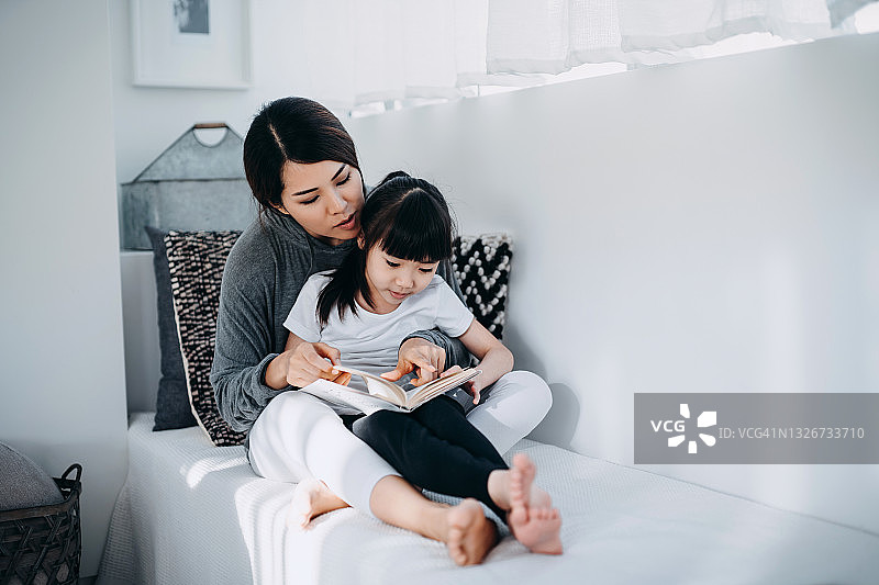 年轻的亚洲妈妈坐在卧室的窗边，给小女儿读书，在家享受家庭 bonding 时光图片素材