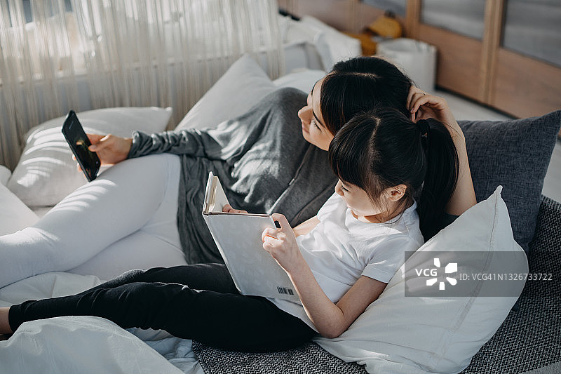 年轻的亚洲母亲和小女儿躺在床上用智能手机看书，在家中享受轻松时光，阳光透过窗户照在她们身上图片素材