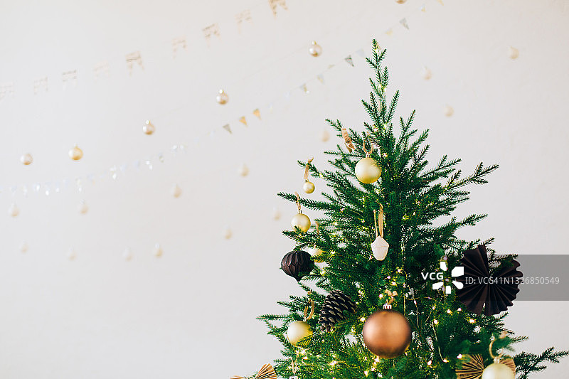 金色古铜色小饰品，纸质装饰品和松果在圣诞树上，靠近纹理白色墙壁和花环。新年庆祝概念。正面图。为您的设计复制空间图片素材