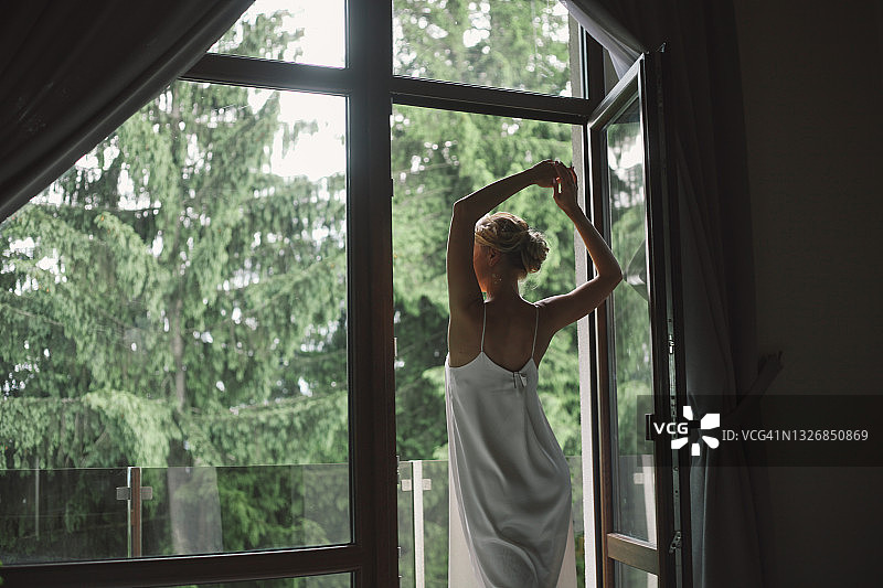 站在阳台上向外看的金发年轻女子，美丽的绿色森林景观，晨间活动概念图片素材